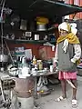 Chaiwala at a stall in Varanasi, a pot of chai is on the stove, and a tray of clean glasses is visible on the table in the background.