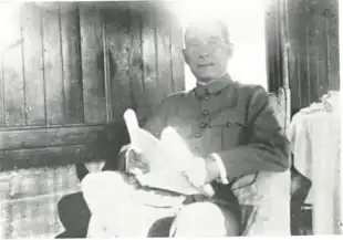 A  black and white photograph of a seated man wearing a military or pseudo-military jacket