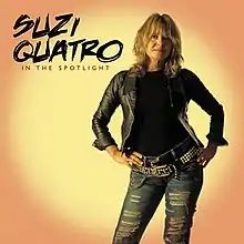 The front cover of Suzi Quatro's album In the Spotlight. Quatro is standing up with her hands on her hips. Behind her is yellow circle on an orange background, simulating the effect of a spotlight. She is wearing a black leather jacket (over a black top) and light blue jeans with horizontal holes ripped in them. She has two black leather belts – a plain one, plus one decorated with yellow studs and yellow chains.