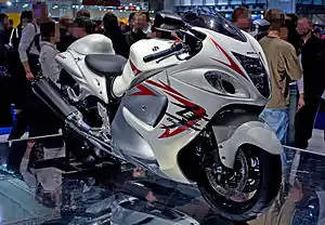 A white motorcycle with fully enclosed bodywork standing upright on a mirrored top platform in a large indoor hall with a crowd of people in the background