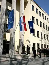 A pale yellow building with square columns with three flags hanging in front and soldiers and dignitaries saluting them.