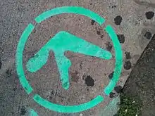 The edge of the pavement on a city street in New York, United States. Green-coloured spray-painted graffiti depicts the Aphex Twin logo inside a dashed outer circle. At the bottom right is roadside greenery.