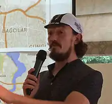  Mike Hall, seated, at a table with a microphone in his hand, before a 2015 race