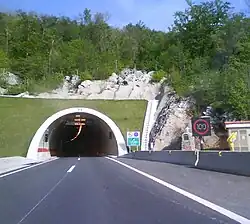 Tunnel portal, variable traffic signs indicating traffic flow direction and speed limit enforced are visible at the tunnel entrance and to the side of the road