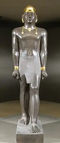 Statue of Taharqa. His name appears on the center of his belt: 𓇿𓉔𓃭𓈎 (tꜣ-h-rw-q, "Taharqa"). The statue is 2.7 meters tall. Taharqa has a striding pose, the arms held tight, and holds the mekes staff. He wears a pleated kilt called shendjyt and on the head the double-uraeus skullcap, possibly signifying the double rule over Nubia and Egypt. (Louvre Museum, color reconstruction of the jewelry through pigment analysis).