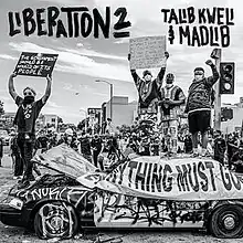 A black and white photo of a crowd in street wear and masks, standing on top of a vandalized car, holding up signs