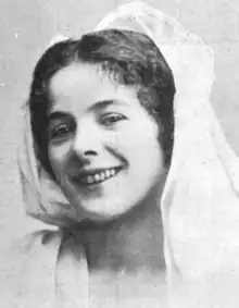 A smiling young white woman with dark center-parted hair with a white scarf draped over her head