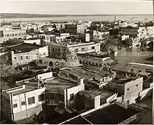 Sepia-toned helicopter view of a city with roads flooded by water and buildings damaged. Another helicopter can be seen near the middle of the image.