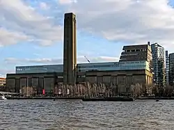 A large oblong brick building with square chimney stack in centre of front face. It stands on the far side of the River Thames, with a curving white foot bridge on the left.