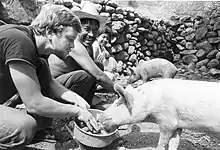 A simple stacked stone wall behind a young Ted Conover, who kneels to feed and coo at a pig while a shirtless Mexican man in a straw cowboy hat and a little Mexican girl kneel nearby, watching Conover and laughing