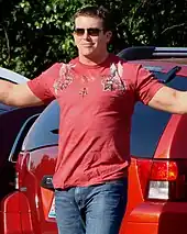 A dark haired young man wearing dark sunglasses poses for cameras with his arms outstretched. He is wearing a red T-shirt, with a pattern on both shoulders and the top part of the chest, and jeans.