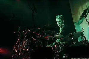 Drummer with crewcut, wearing black T-shirt, intently plays his large drum set in the spotlight.