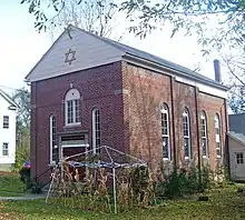 A brick building with a small gold-on-black sign saying "Congregation Beth David" above the door, round-arched windows and a small structure in front made of withered cornstalks on a metal frame