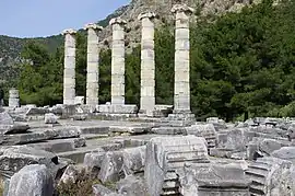 Five upright columns surrounded by ruined stonework, in front of a hill.