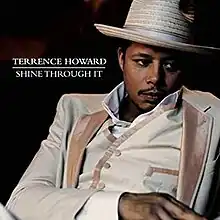 A photo of the artist leaning back in a chair, wearing a white suit and fedora, in front of a black background.