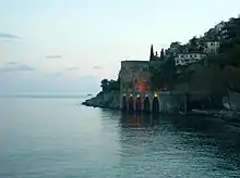 A hill populated with houses slants down into a blue-green sea below a stone dock with five arches. A stone wall extends along the sea from the dock to the lower right.