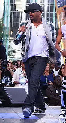 An Afro-American man wearing a white shirt, a black cap and sunglasses is walking onto a stage while holding a microphone.