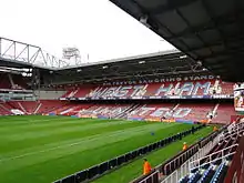 A stand from a football stadium with laid out turf and two adjacent goals in the foreground. On the claret stands are the words written in sky blue capitals "Sir Trevor Brooking stand" on top of the larger words "West Ham United". There are men in orange jackets scattered around the stadium.