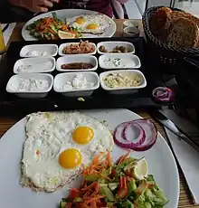 Plates with fried eggs, green salad, and onions; basket of bread; smaller dishes filled with variety of sides.