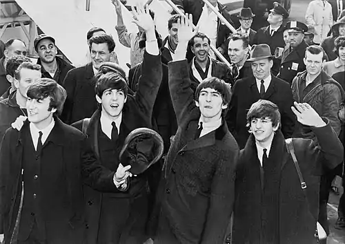 The Beatles are standing in front of a crowd of people at the bottom of an aeroplane staircase.