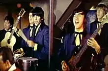 A band consisting of five men wearing royal blue suits, white shirts and black ties, is performing in front of a beige-and-brown wall. Two men (far left, fourth from left) are playing guitars, while two others (middle, far right) are playing percussion instruments. Four band members have dark brown or black hair; the man on the far right is blond. A pole in the foreground separates the three men on the left from the two on the right.