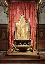 An old gilded wooden chair on a raised platform in front of a red curtain.