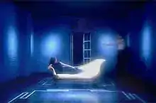  In a room filled with blue light, a woman reclines on an antique couch, and a man standing nearby has one arm outstretched.