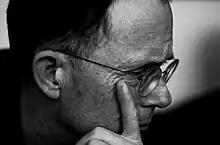 Profile of a bespectacled middle-aged white man. He is looking to the right, and his left index finger rests on his right cheek slightly below the eye.