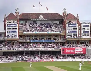 A three tiered Victorian red-brick building with flags flying on the roof and a clock at the top. There are large numbers of spectators watching a cricket match.