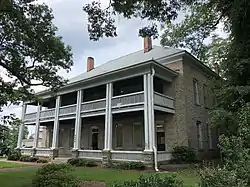 A two story granite block structure with a hipped roof and two-story wooden veranda, seen from an angle in front.