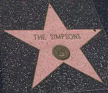 A coral-pink terrazzo five-point star inlaid into a charcoal-colored terrazzo background. In the upper portion of the pink star field, the text "The Simpsons" is inscripted. Below the inscription, in the lower half of the star field, there is a round emblem with an image of a television in it.