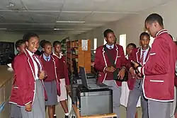 8 students in school uniforms stand talking to each other in a classroom or library. They are wearing white or blue button-down shirts, a red and blue or red and white striped tie, a red vest, and either a grey skirt or trousers. Between the students are laptops sitting atop printers.