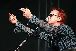 A color photograph of a man, facing him from his left side, on stage at a concert, singing into a microphone and pointing his fingers at the crowd.