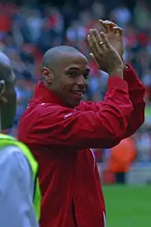 Thierry Henry, wearing a red shirt with white long sleeves and shorts with a number 12 and Nike logo on the left-leg side, applauds.