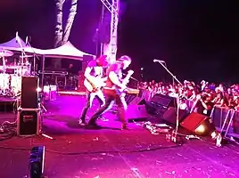 Side of stage shot of a band playing. Two members are shown in right profile both are playing guitars. Other stage equipment is visible around them. The audience is to the right and below stage level.