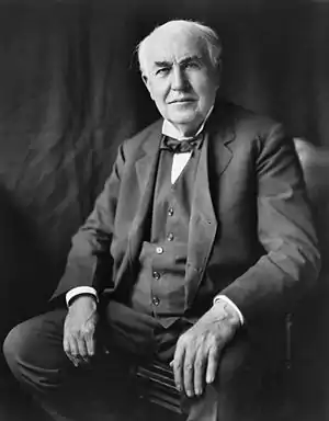 An aged man sitting in a chair. His hands are at his hips.