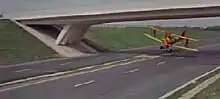 A yellow and red biplane flies underneath a deserted motorway bridge.