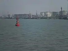 A red port-side buoy with an oil wharf and tankers in the background