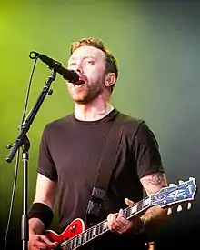 A caucasian man wearing a black shirt. The man is holding a red guitar while singing in front of a mircophone