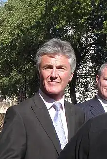 A middle-aged man with grey hair wearing a suit looks at the camera with a tree backdrop