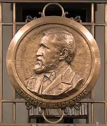 a circular bronze plaque has a raised impression of the bust of a man who is facing to the left.  The place is located on bronze railings.