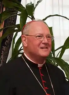 A bespectacled man wearing liturgical vestments and a chain around his neck attached to a pectoral cross that is not visible in photograph facing right