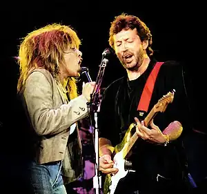 Turner and Clapton, on stage, sharing a microphone stand, singing.