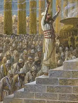 In the foreground, a bearded man dressed in an impressive white robe and head-dress raises his hand to heaven.  Behind him, a large crowd bows in prayer.