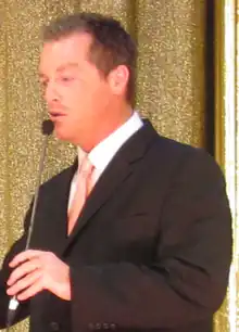 A man with dark hair wearing, including a black suite with a pink tie.