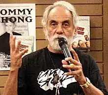  A well-tanned man with white hair and a white beard holds a microphone in front of his face. On his left wrist, he wears a heavy silver-colored watch; with his right hand, he is gesturing. On the wall behind him are two signs: one bears the name "Tommy Chong".