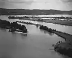 A black and white aerial view of a bay on a lake with a small fishing boat with two men aboard in the foreground.