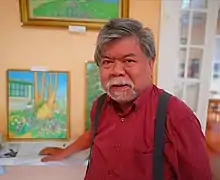 A man in a red shirt and suspenders stands next to several paintings on the wall of an art gallery.