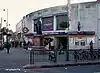 A grey-bricked building with a rectangular, dark blue sign reading "TOOTING BROADWAY STATION" in white letters all under a grey sky