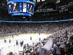  An arena set up for ice hockey. Players skate towards center ice, while a larger crowd is waiving white towels. Over head is a jumbo-tron with an extreme close-up of a player looking seriously at the camera, above and below the picture is the word "Believe" in white lights with a blue light background.